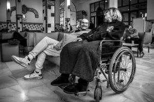 Fotografía en blanco y negro de una mujer en silla de ruedas conversando con un hombre en una sala.